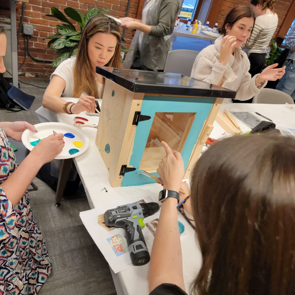 Participants focus on painting a Little Free Library door with bright orange during a collaborative team building event. The hands-on activity fosters creativity and teamwork as teams work together to build and decorate the library for community use.