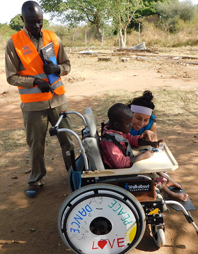 A young boy receiving a decorated wheelchair during the Wheels Around the World team building event, accompanied by two adults who are assisting him outdoors.