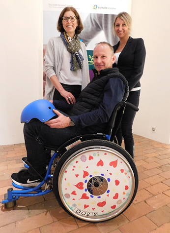 Two team members pose next to a man seated in a decorated wheelchair featuring heart designs on the wheel cover, created during the Wheels Around the World team building event.