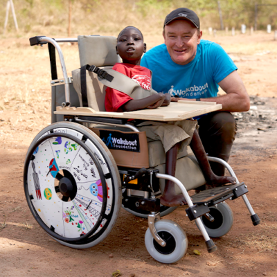 A young boy sits in a newly donated wheelchair, customized with a decorated wheel safety cover, next to a smiling volunteer from the Walkabout Foundation during the Wheels Around the World team building event.