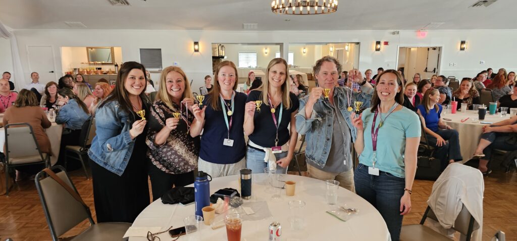 Group of participants celebrating at a team building event, holding small trophies while posing in front of an audience in a large room.