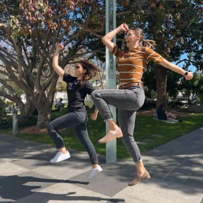 Two women energetically jump in the air during a team building activity outdoors, with trees in the background. Both are striking playful poses mid-jump, adding a sense of fun and excitement to the moment.