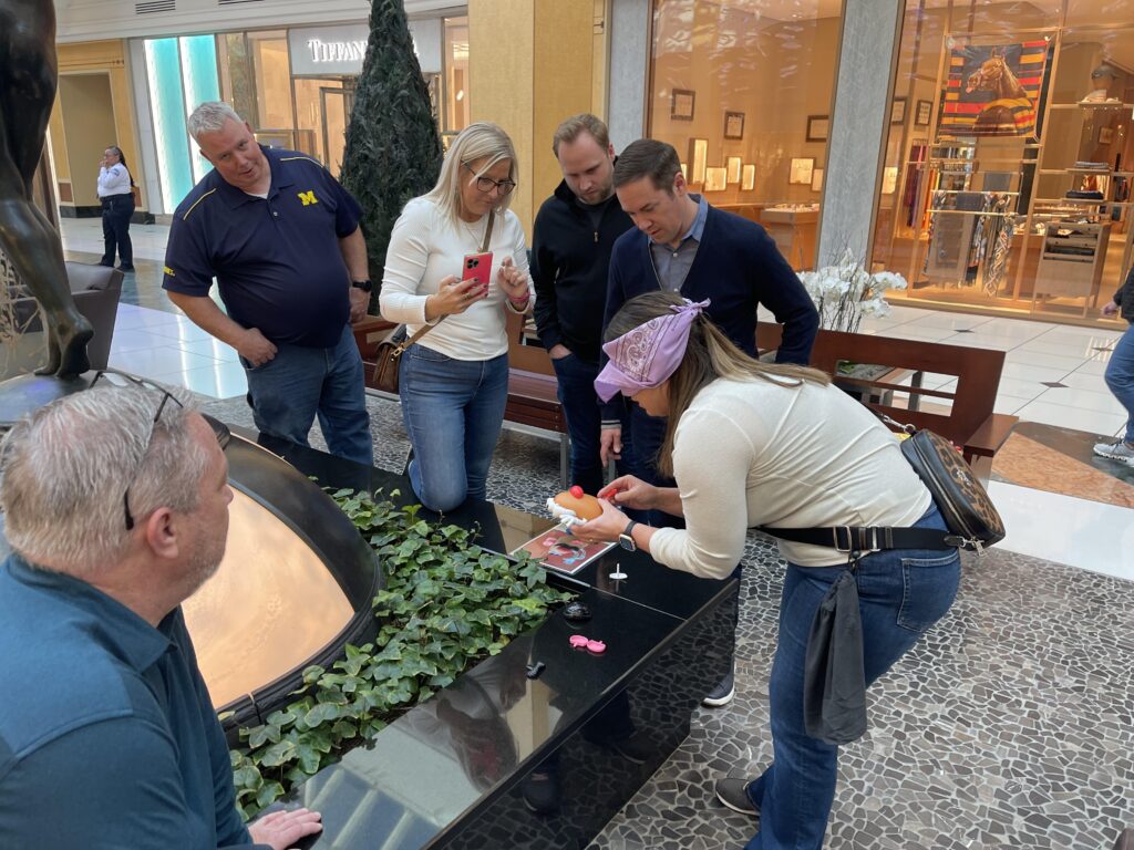 Team building participants gather around a table in a mall as one blindfolded member engages in a challenge involving assembling a toy.