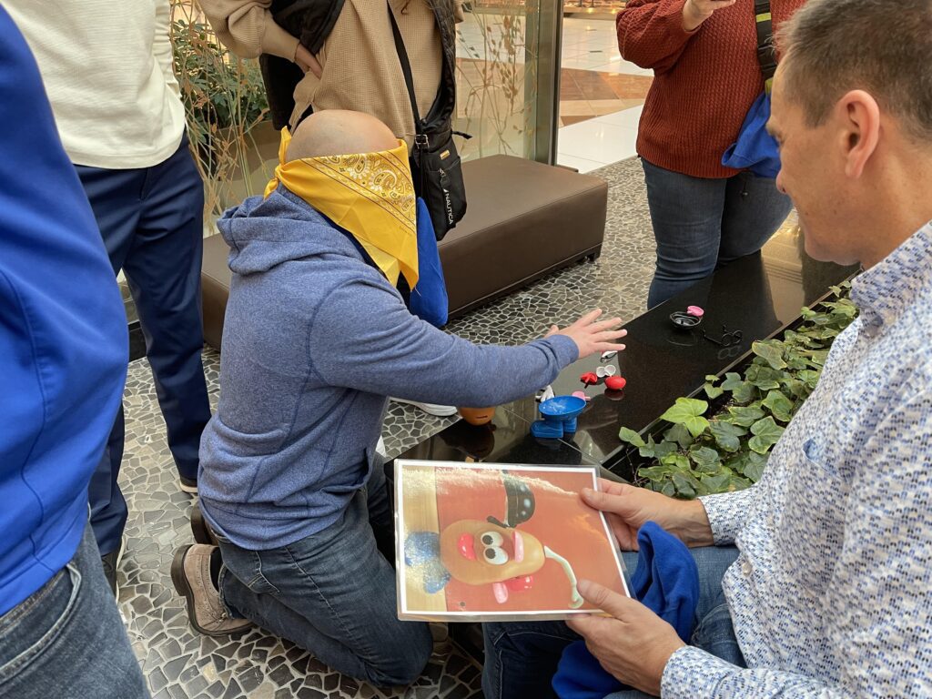 A blindfolded participant engages in a team building challenge, guided by another team member holding a picture as they work together in a collaborative indoor activity.