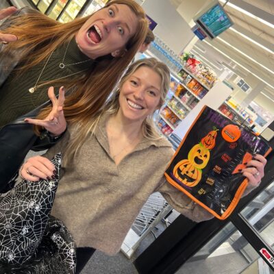 Two women are standing in what appears to be a grocery store, smiling and posing for the camera. The woman on the left has long red hair, is wearing a green sweater, and is making a playful spooky face with her hands raised as if pretending to be a monster. The woman on the right is smiling brightly and holding up a Halloween-themed reusable shopping bag with a jack-o'-lantern design. The scene is part of a Halloween-themed team building challenge.