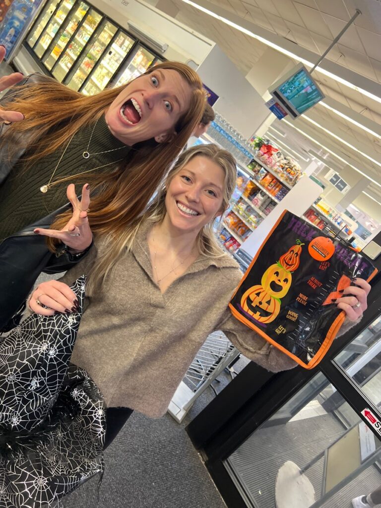 Two women are standing in what appears to be a grocery store, smiling and posing for the camera. The woman on the left has long red hair, is wearing a green sweater, and is making a playful spooky face with her hands raised as if pretending to be a monster. The woman on the right is smiling brightly and holding up a Halloween-themed reusable shopping bag with a jack-o'-lantern design. The scene is part of a Halloween-themed team building challenge.