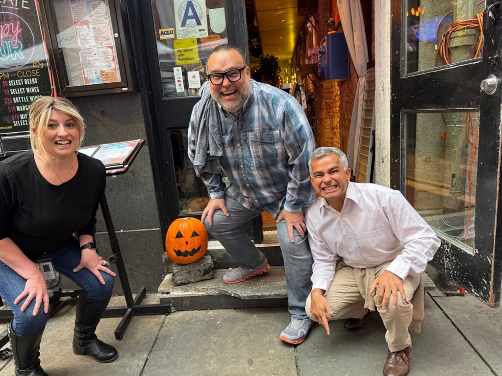 Three people are crouching and smiling outside a building with a carved pumpkin placed on the steps beside them. The person on the left is a woman with blonde hair, wearing a black top and jeans. In the middle is a man wearing glasses, a plaid shirt, and gray pants. The person on the right is another man dressed in a white shirt and khaki pants. The group is engaging in a Halloween-themed team building activity.