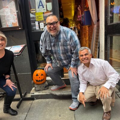 Three people are crouching and smiling outside a building with a carved pumpkin placed on the steps beside them. The person on the left is a woman with blonde hair, wearing a black top and jeans. In the middle is a man wearing glasses, a plaid shirt, and gray pants. The person on the right is another man dressed in a white shirt and khaki pants. The group is engaging in a Halloween-themed team building activity.