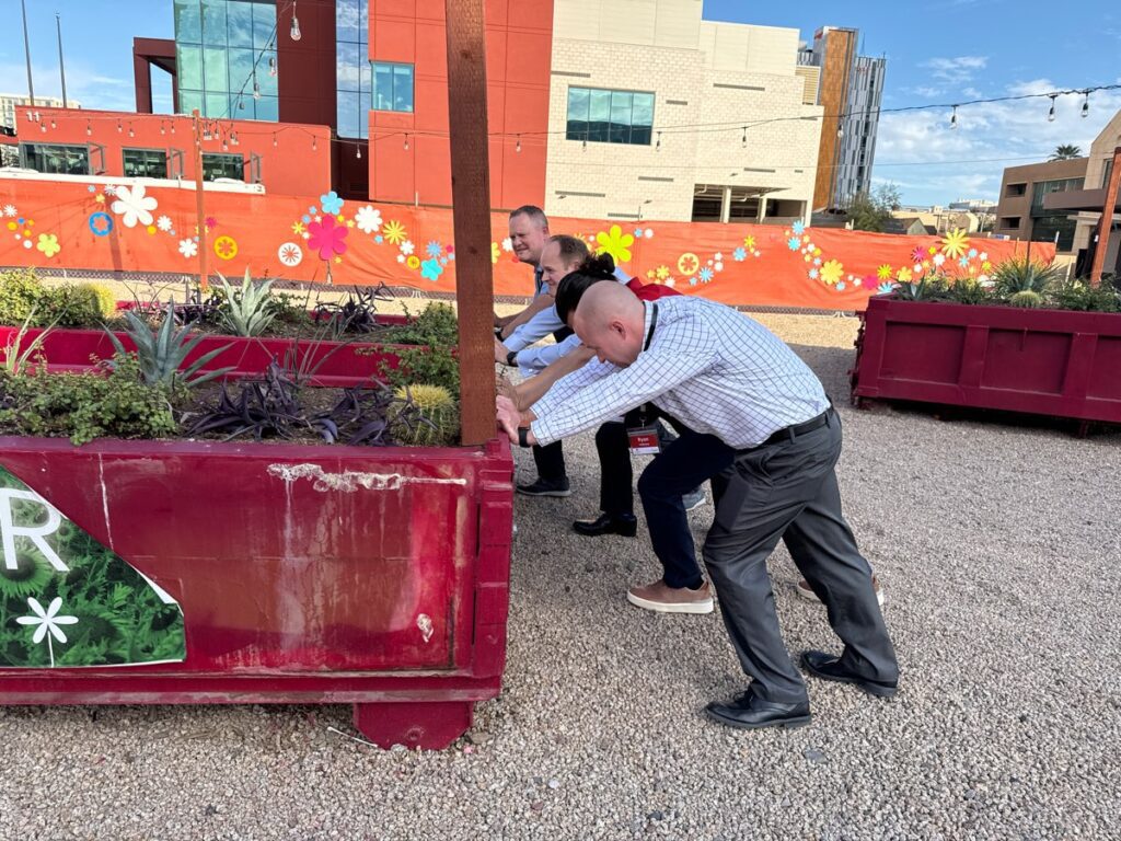 Three participants in a team building activity work together to push a large planter in an outdoor space.