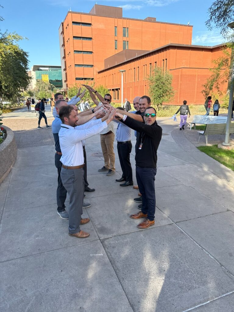 A group of men forming an arch with their arms during a team building exercise outdoors, with a red brick building in the background.