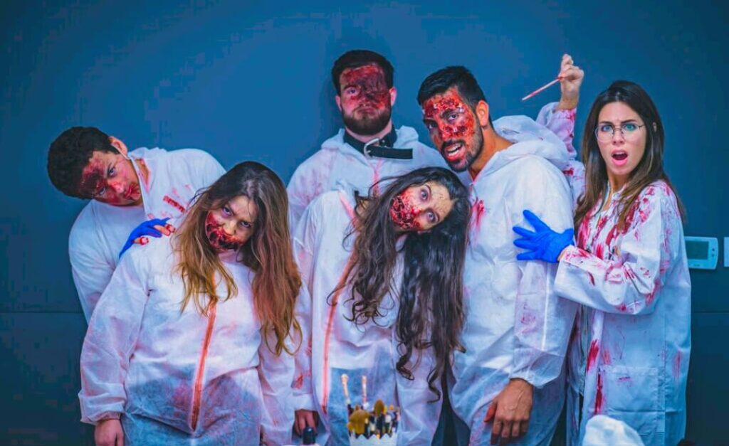 A group of participants dressed as zombies in white blood-stained protective suits poses for a photo during a zombie-themed team building event. Their faces are covered in detailed zombie makeup, with expressions of shock, confusion, and eerie detachment, adding to the spooky atmosphere of the activity.