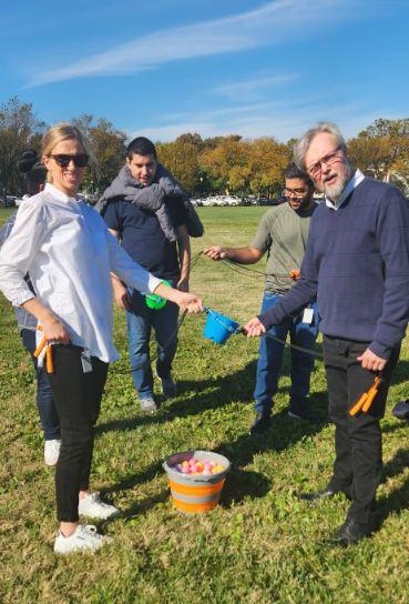 A group of participants working together in a team building activity outdoors. They are holding ropes to lift and transport a small bucket filled with colorful items, practicing collaboration and communication.