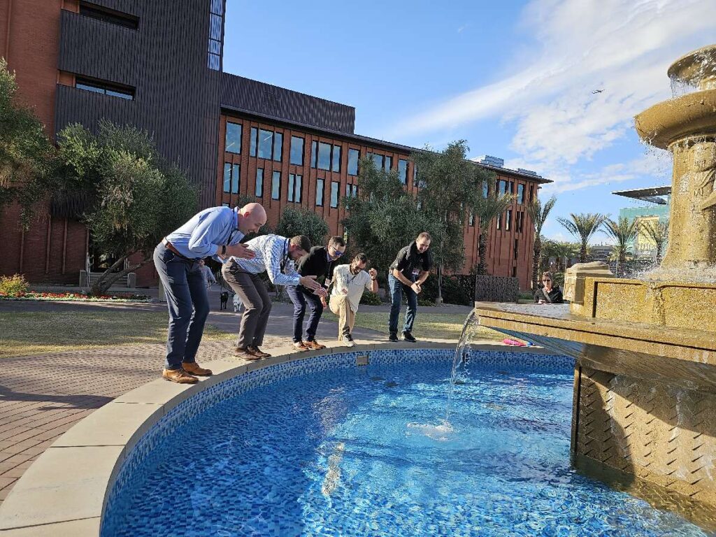 Group of five team building participants crouched near a water fountain, preparing to take action during an outdoor team building activity.