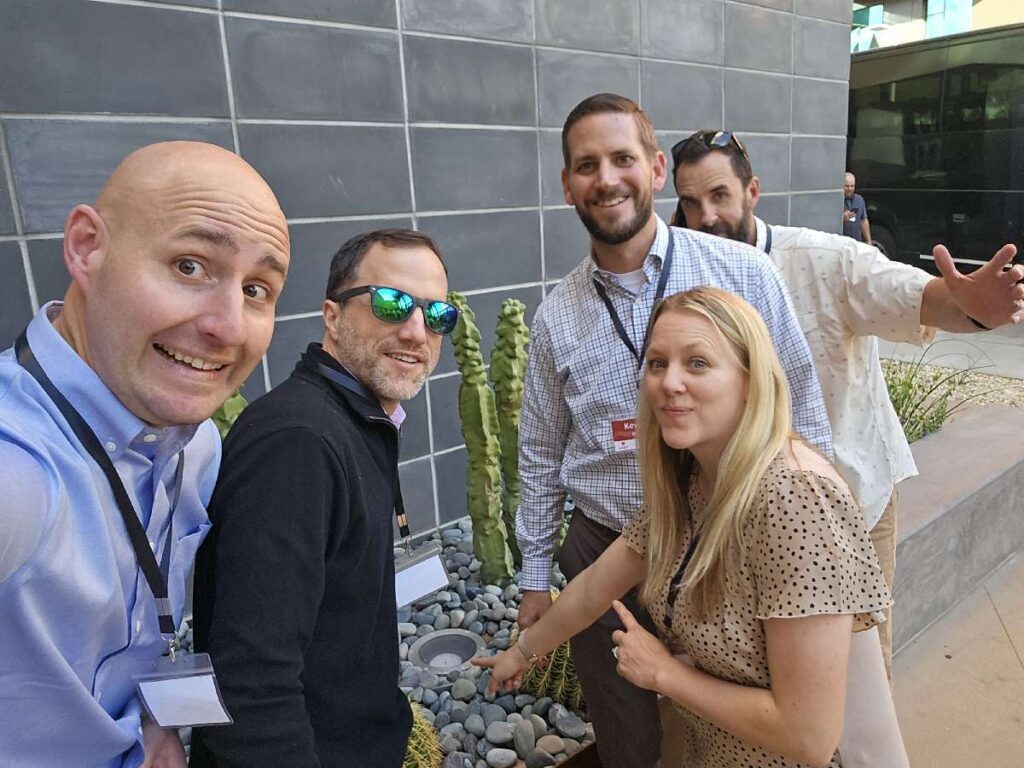 Group of five team building participants posing next to a small garden display, smiling and pointing at an object during an outdoor team activity.