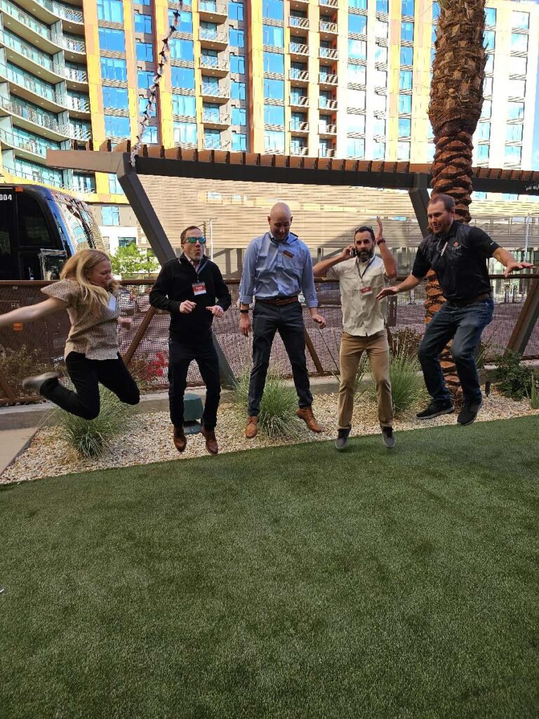 Five participants jumping in the air on a grass field during an outdoor team building event, with buildings in the background.