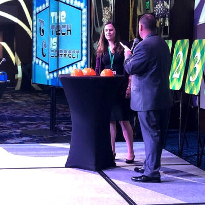 A host engages a contestant in the Play it Forward game show event on stage, with a screen in the background displaying the phrase 'The Tech is Correct.' The stage is set for a trivia challenge, and the contestant stands next to a table with orange buttons used in the game. Numbered boards are visible on the side, representing various challenges in this charitable team building activity.