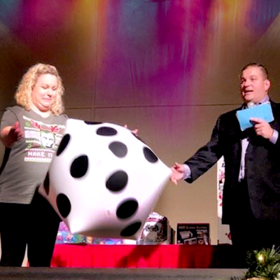 A contestant rolls a large inflatable die on stage during the Play it Forward charity team building game show event, while the host watches and holds game cards.