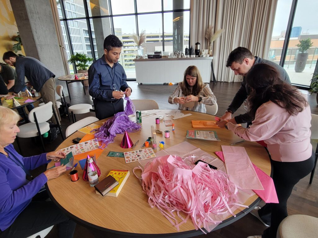 A group of people is gathered around a table, engaged in a crafting activity. The table is covered with materials like colorful paper, party hats, stickers, and pink streamers as they work on assembling birthday kits to be donated as a result of this charitable team building program.