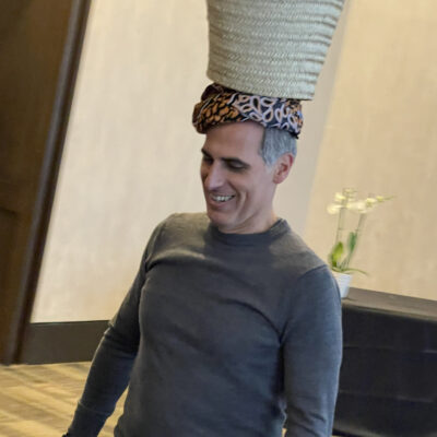 A smiling man participates in a water walk challenge by balancing a basket on his head during a Clean Water Connection team building event.