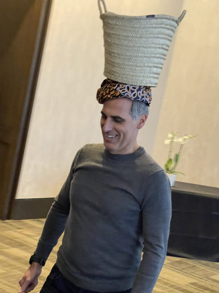 A smiling man participates in a water walk challenge by balancing a basket on his head during a Clean Water Connection team building event.