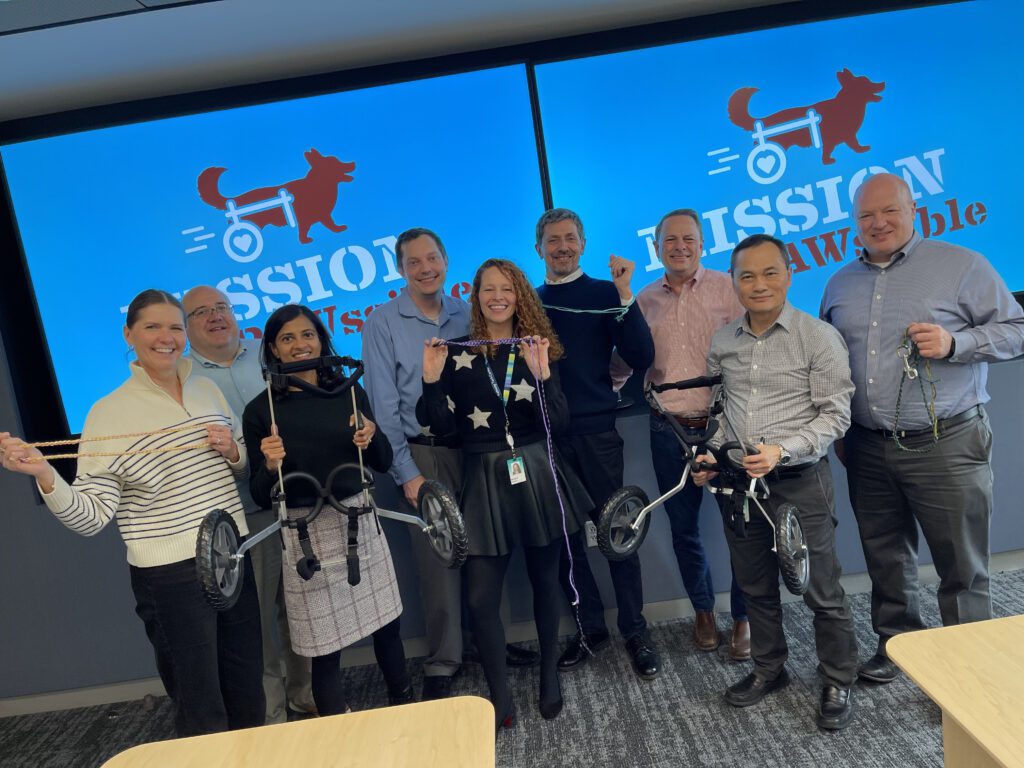 Group of smiling participants posing with assembled pet wheelchairs, standing in front of a screen displaying 