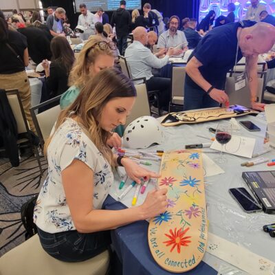 A woman seated at a table carefully decorating a skateboard with colorful designs of flowers and inspirational text. The surrounding table is filled with art supplies, a helmet, and other tools. Behind her, other participants are also engaged in the creative process during a team building event focused on assembling and decorating skateboards for a charitable cause.