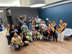 A group of participants proudly displaying their decorated skateboards and helmets after a successful team building event. The group, consisting of around 20 individuals, poses with their custom creations in a brightly lit modern office space. The skateboards and helmets are painted in various colorful designs, showcasing the team's creativity and collaboration. The event celebrates teamwork and artistic expression, bringing everyone together for a charitable cause.