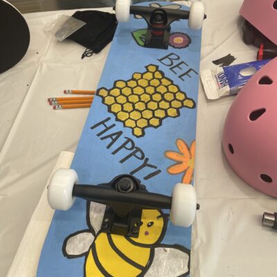 A custom-decorated skateboard featuring a cheerful design with a honeycomb, a bee, and the phrase 'BEE HAPPY!' The board has white wheels, and next to it on the table are some pencils, a pink helmet, and various crafting supplies. The skateboard is part of a creative team building event where participants assemble and decorate skateboards for a charitable cause.