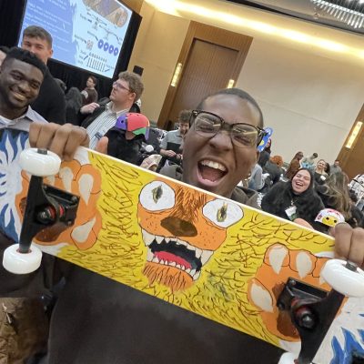 Excited participant proudly holds up a creatively designed skateboard during the Just Roll With It team building event, where teams build and decorate skateboards for charity.