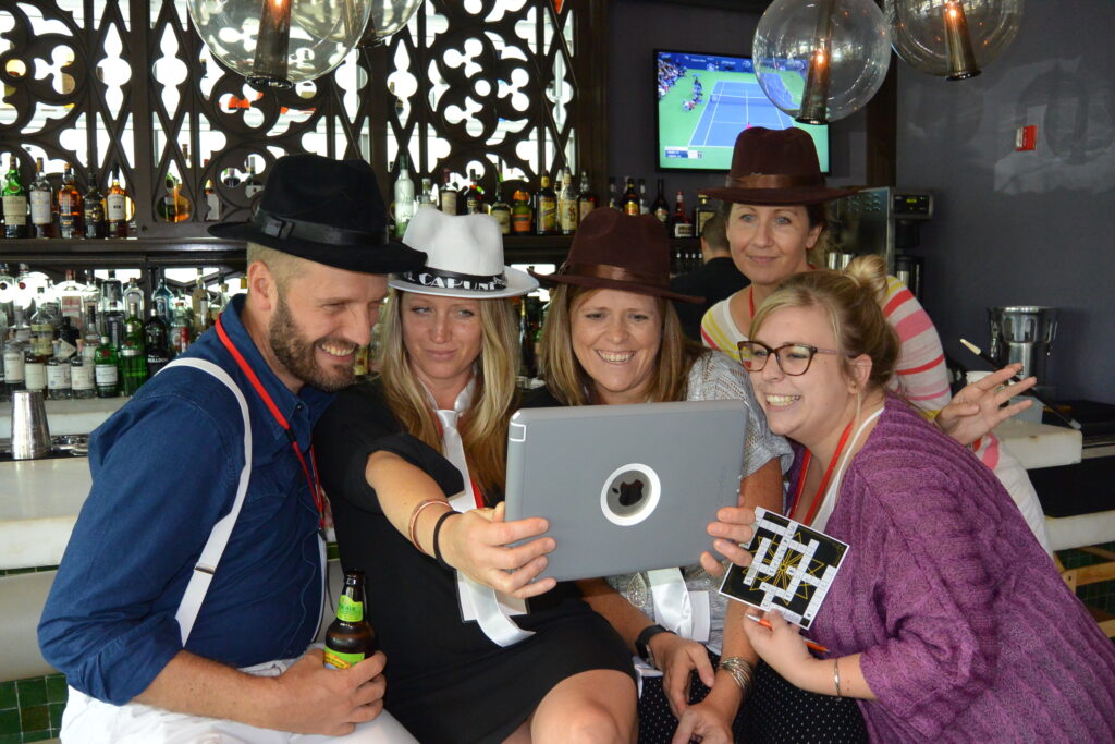 Team members wearing detective hats gathered around a tablet during the Sleuth: The Murder Mystery Scavenger Hunt team building activity, smiling and analyzing clues together