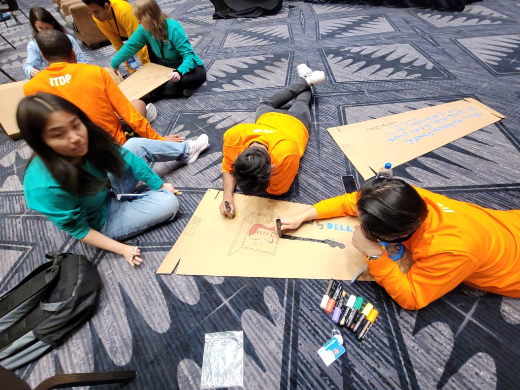 Team members lying on the floor, focused on decorating their cardboard guitar cases with colorful markers during the Charity Guitar Build team building event.