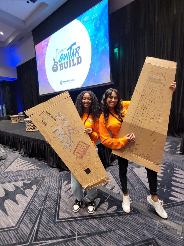 Two participants holding custom-decorated cardboard guitar cases at the Charity Guitar Build team building event, supporting music education for aspiring musicians.