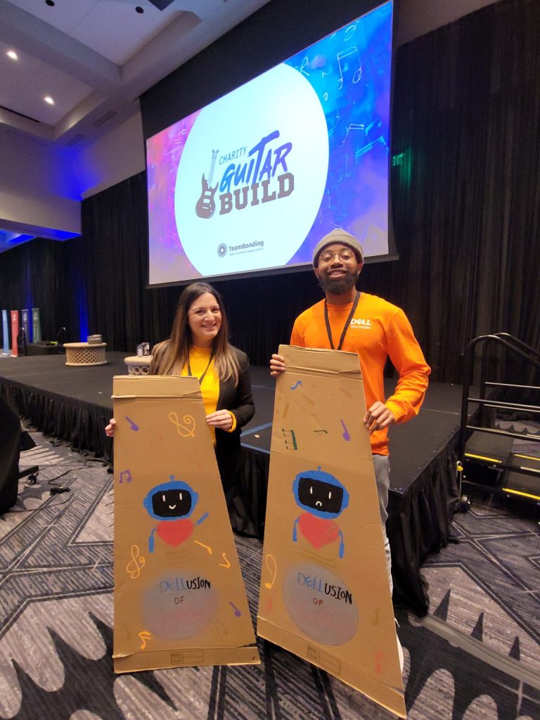 Two team members proudly display their decorated cardboard guitar cases during the Charity Guitar Build team building event, showcasing their creativity and enthusiasm.