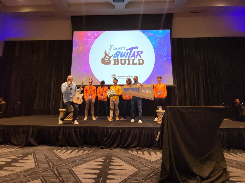 A team on stage at the Charity Guitar Build team building event proudly presents their finished guitar and decorated case, celebrating teamwork and creativity for a charitable cause.