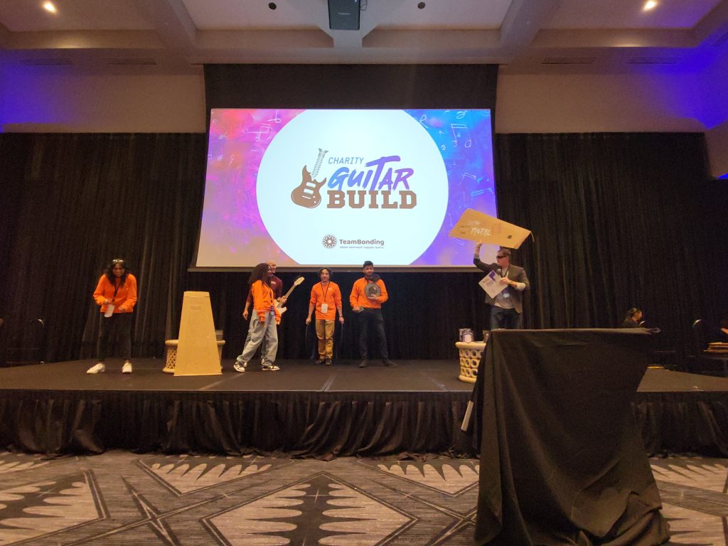 Participants on stage presenting their decorated guitar case at the Charity Guitar Build event, showcasing creativity and teamwork in support of aspiring musicians.