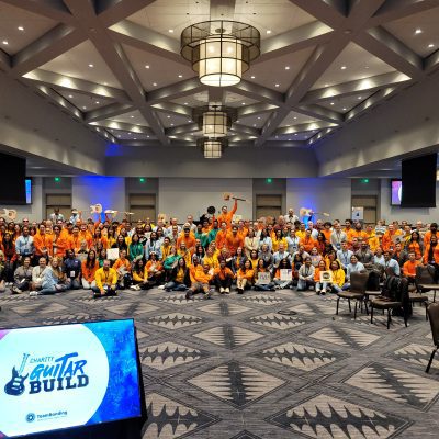 Large group photo at the Charity Guitar Build team building event, with participants holding guitars and celebrating teamwork and community impact.