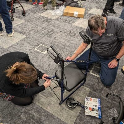 Two participants are kneeling on the floor, carefully assembling a rollator walker by attaching its handles and frame components during a charitable team building activity.