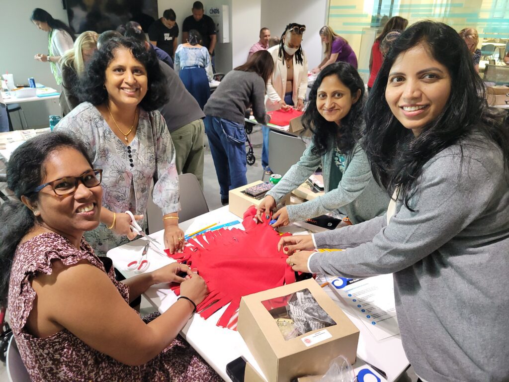 A group of four women smiling while working together on a no-sew fleece blanket, tying knots during a charitable team building event.