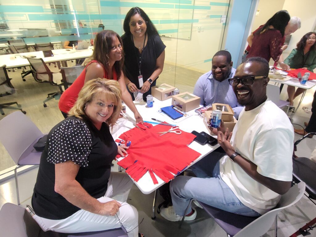 Five smiling participants sit around a table in a brightly lit room, engaged in assembling items for the Rollator Walker Build team building program. On the table are red fabric pieces, scissors, and small boxes, as they work on decorations and assembling parts. The group looks cheerful as they work together.