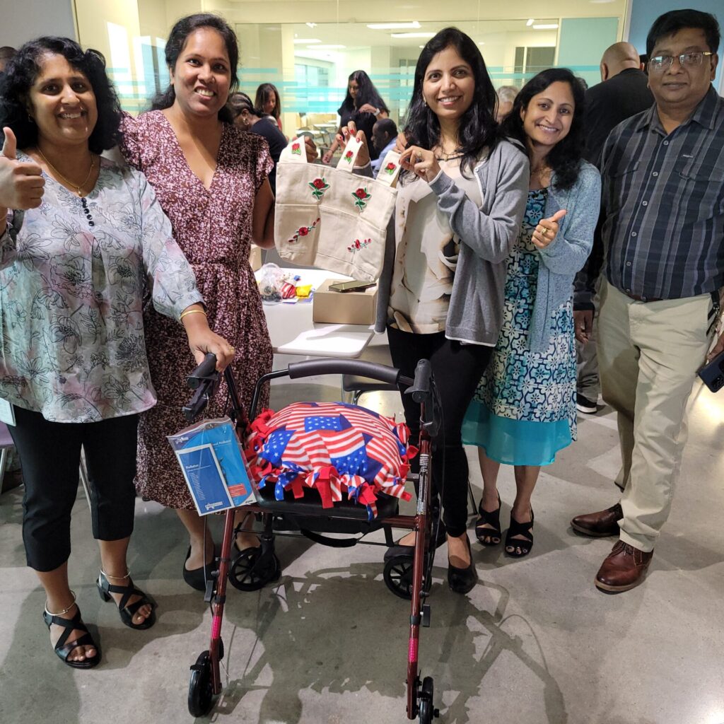 A group of five individuals, smiling and showing a thumbs up, stands around a rollator walker they have built. The walker features an American flag-themed cushion attached to it. One of the women is holding up a canvas tote bag decorated with colorful designs. Various supplies and items are visible on the table behind them, contributing to the Rollator Walker Build team building event.