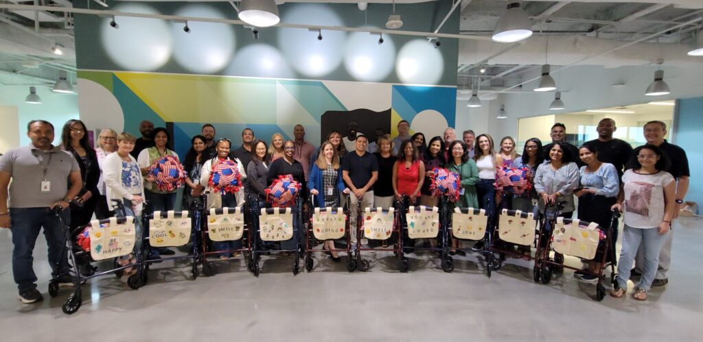 A group of participants stand behind a row of assembled rollator walkers, each adorned with decorated canvas bags and colorful fleece blankets, showcasing their completed project accomplished during a charitable team building program.