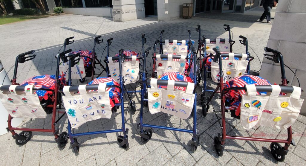 Group of assembled rollator walkers, decorated with personalized tote bags and American flag-themed pillows, lined up outdoors to showcase the result of a corporate charitable team building event.