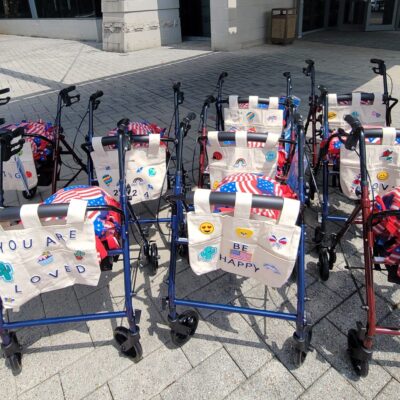 Group of assembled rollator walkers, decorated with personalized tote bags and American flag-themed pillows, lined up outdoors to showcase the result of a corporate charitable team building event.