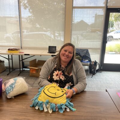 Smiling woman sits at a table holding a brightly colored no-sew pillow with a smiley face design, part of the Rollator Walker Build team building activity.