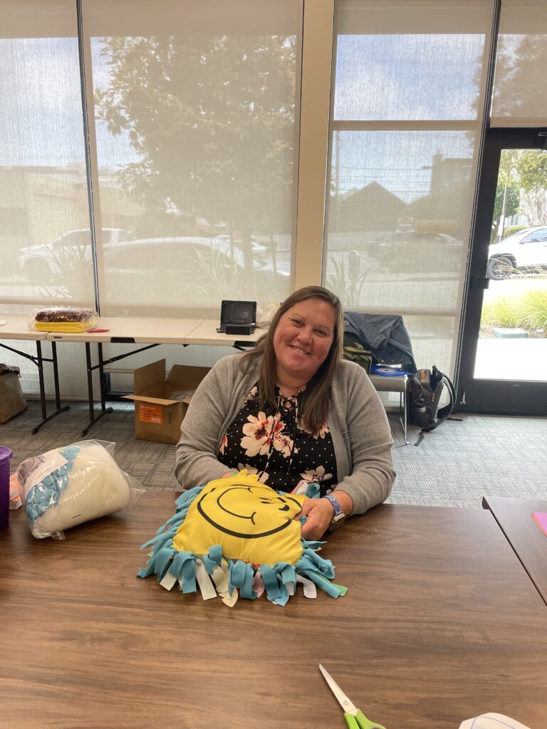 Smiling woman sits at a table holding a brightly colored no-sew pillow with a smiley face design, part of the Rollator Walker Build team building activity.