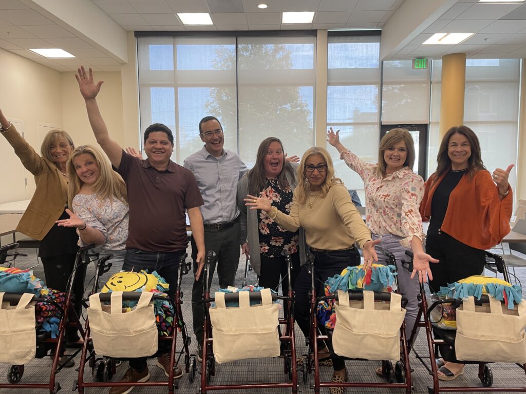 diverse group of eight adults joyfully posing behind their assembled rollator walkers, each adorned with a colorful fleece and a personalized accessory bag, showcasing their collaborative effort in a well-lit room.