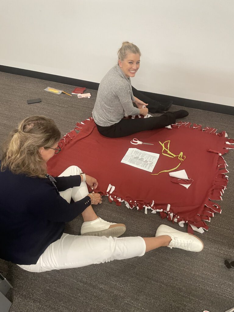 Two women sit on the floor, tying knots on the edges of a red fleece blanket, collaborating on a no-sew team building project.