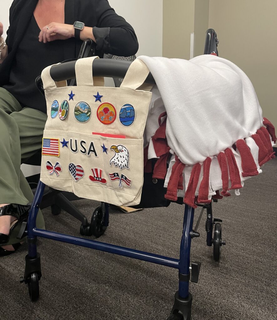 A rollator walker is decorated with a canvas bag displaying various USA-themed patches and a white and red fleece blanket draped over the seat.