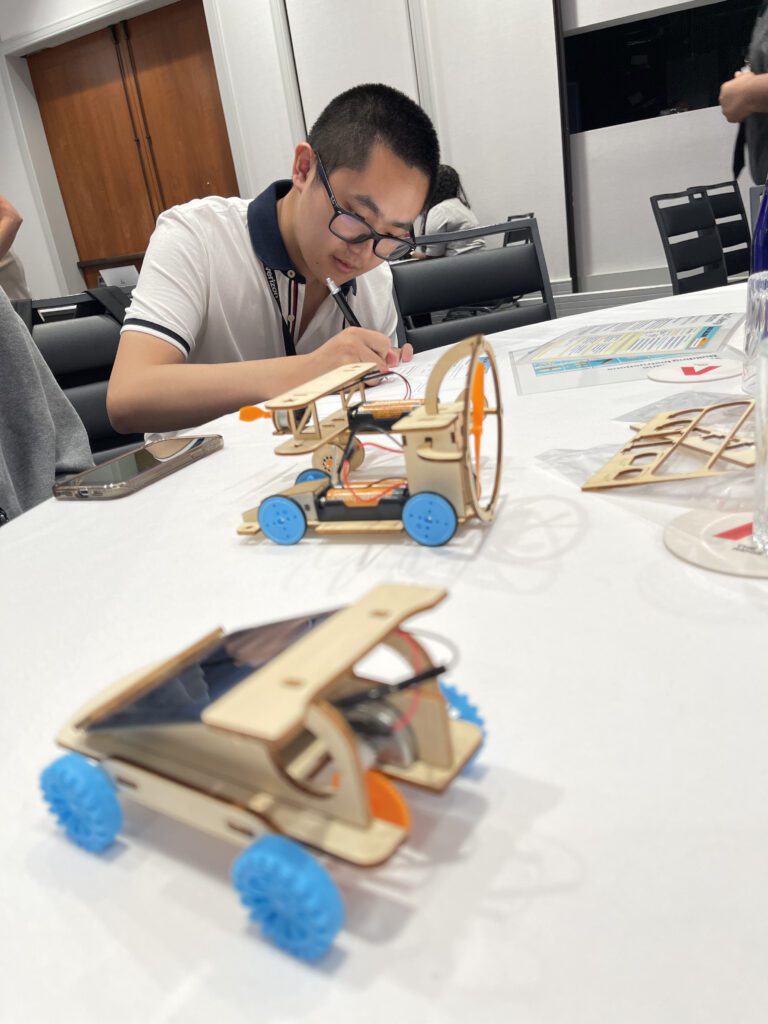 A participant focused on assembling a miniature car from a STEM kit during the STEM Speedway team building activity, with completed wooden model cars on the table, highlighting precision, creativity, and hands-on learning.