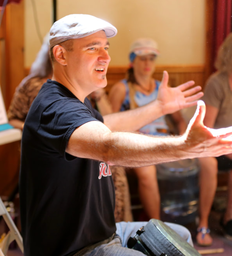 Josh Robinson passionately engages the audience during an interactive drumming session. Wearing a casual black shirt and a light-colored cap, he extends his arms, leading the group in rhythm and connection, creating an immersive and energizing experience.